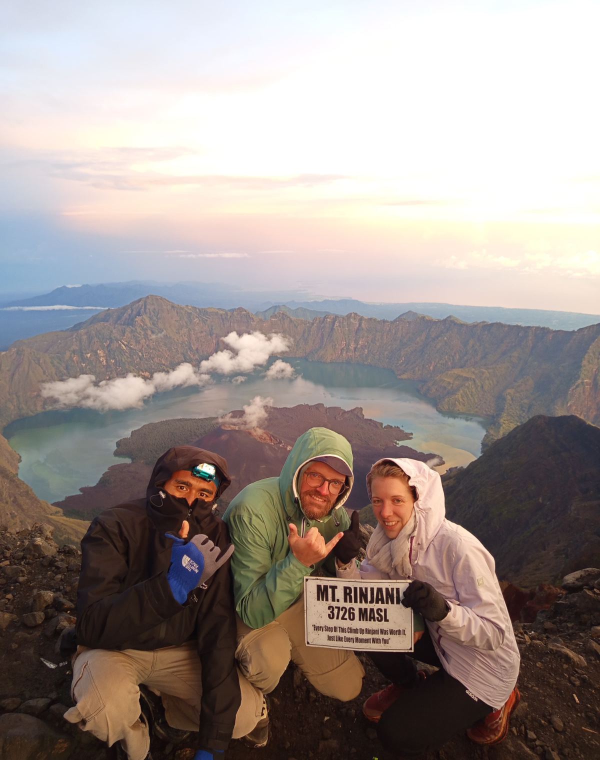 Trekkers holding the Mt Rinjani 3,726 m summit sign at sunrise, with the crater lake far below